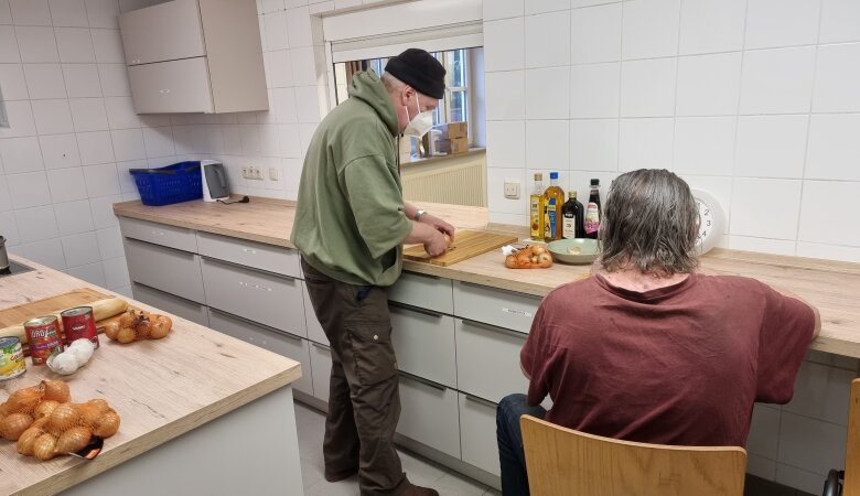 Zwei Männer bereiten Essen in der Küche zu, dabei steht ein Mann am Schneidebrett und ein Mann sitz